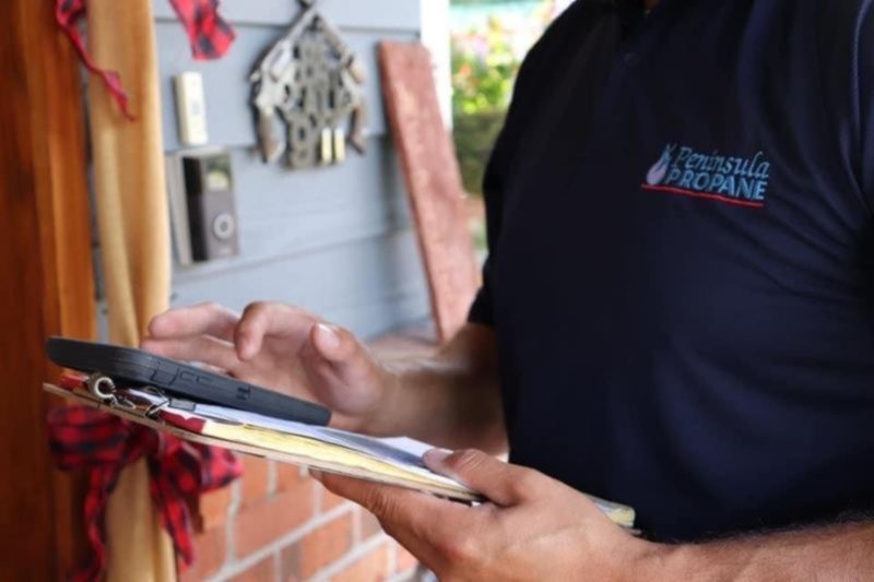 A Peninsula Propane driver in a company polo at a customer's front door with delivery paperwork.